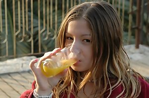 a young woman drinking a glass of orange juice from a glass cup in front of her face and a fence in the background with a metal fence in the and a fence in the foreground and trees in the foreground
